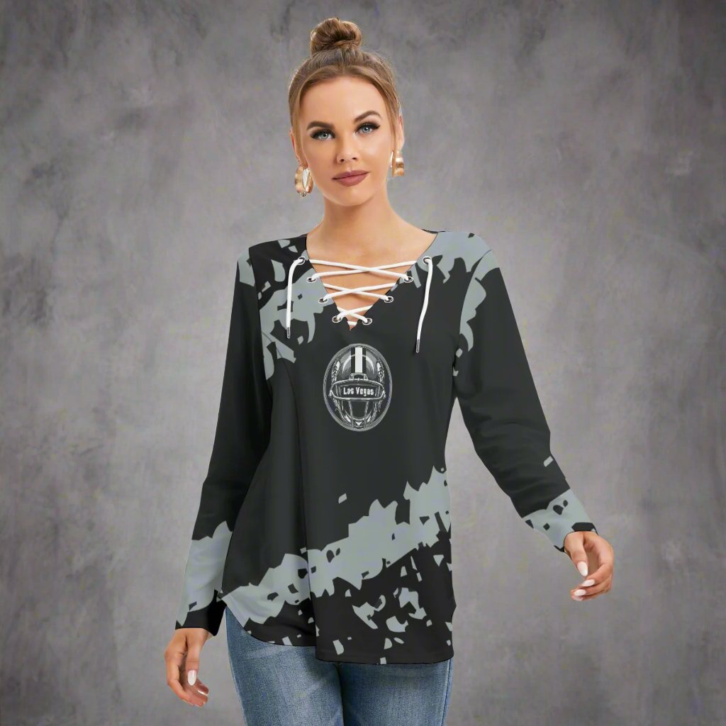 A woman wearing the Las Vegas Women's Shoestring Sweatshirt, a black and gray splattered long-sleeve top with a circular graphic logo and neckline tie, paired with blue jeans, stands against a white background.