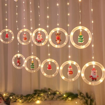 The warm yellow glow of the Christmas LED Light Strings illuminates holiday decorations featuring Santa, reindeer, Christmas trees, and stars against a white curtain backdrop. These festive lights are conveniently powered by USB, adding charm to any setting.