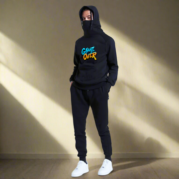 A person wearing a "Game Over" Men's Black Turtleneck Hoodie with Pockets, matching pants, and white sneakers stands on a plain background.